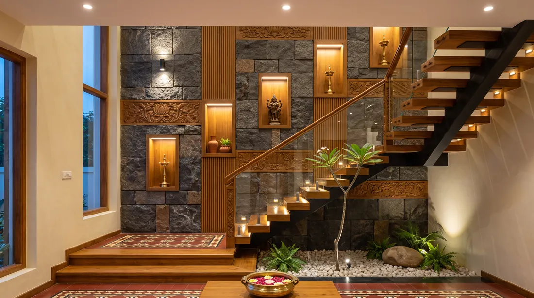 A double-height staircase with open teak treads beside a granite wall with carved teak panels and lit niches holding brass lamps